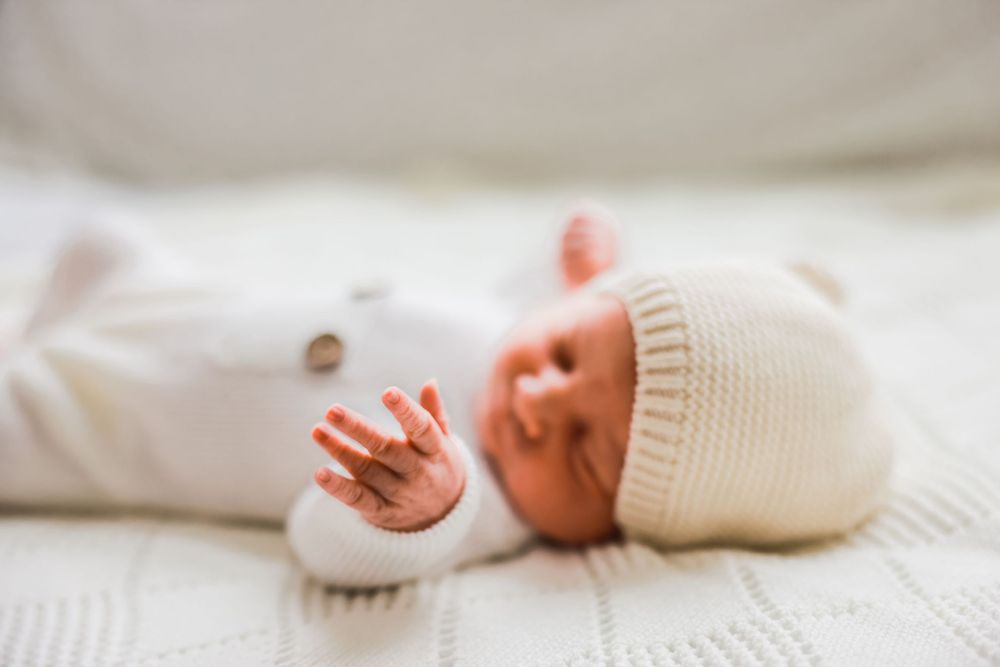 Newborn baby wearing white knit hat and outfit lying on white blanket.