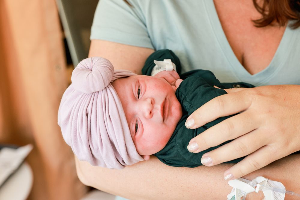 Newborn baby wearing pink head wrap and dark clothing rests peacefully.
