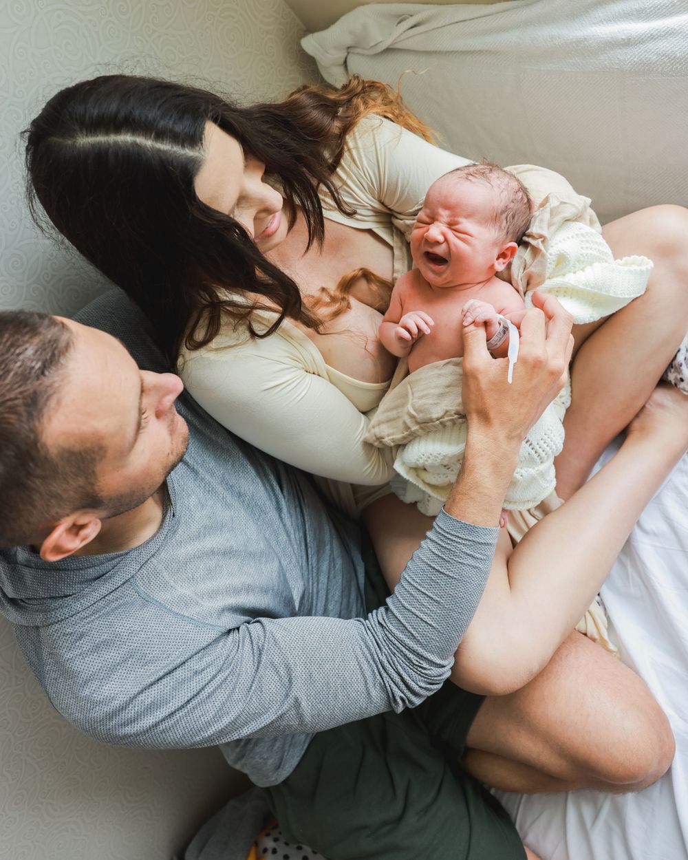 Candid moment of parents lying on bed with their crying newborn baby.