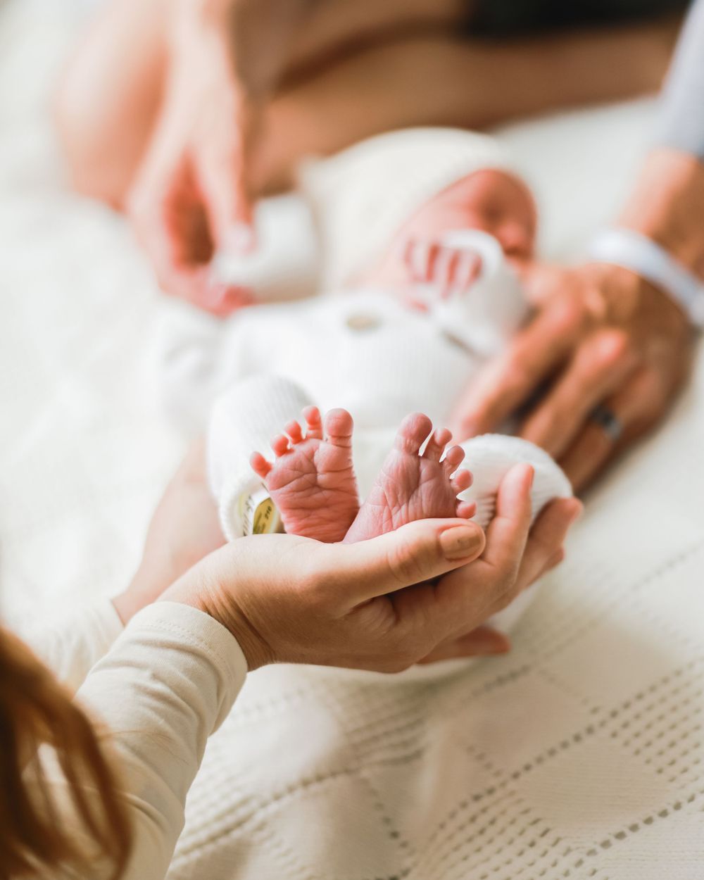 Gentle hands holding tiny newborn feet against white textured bedding.