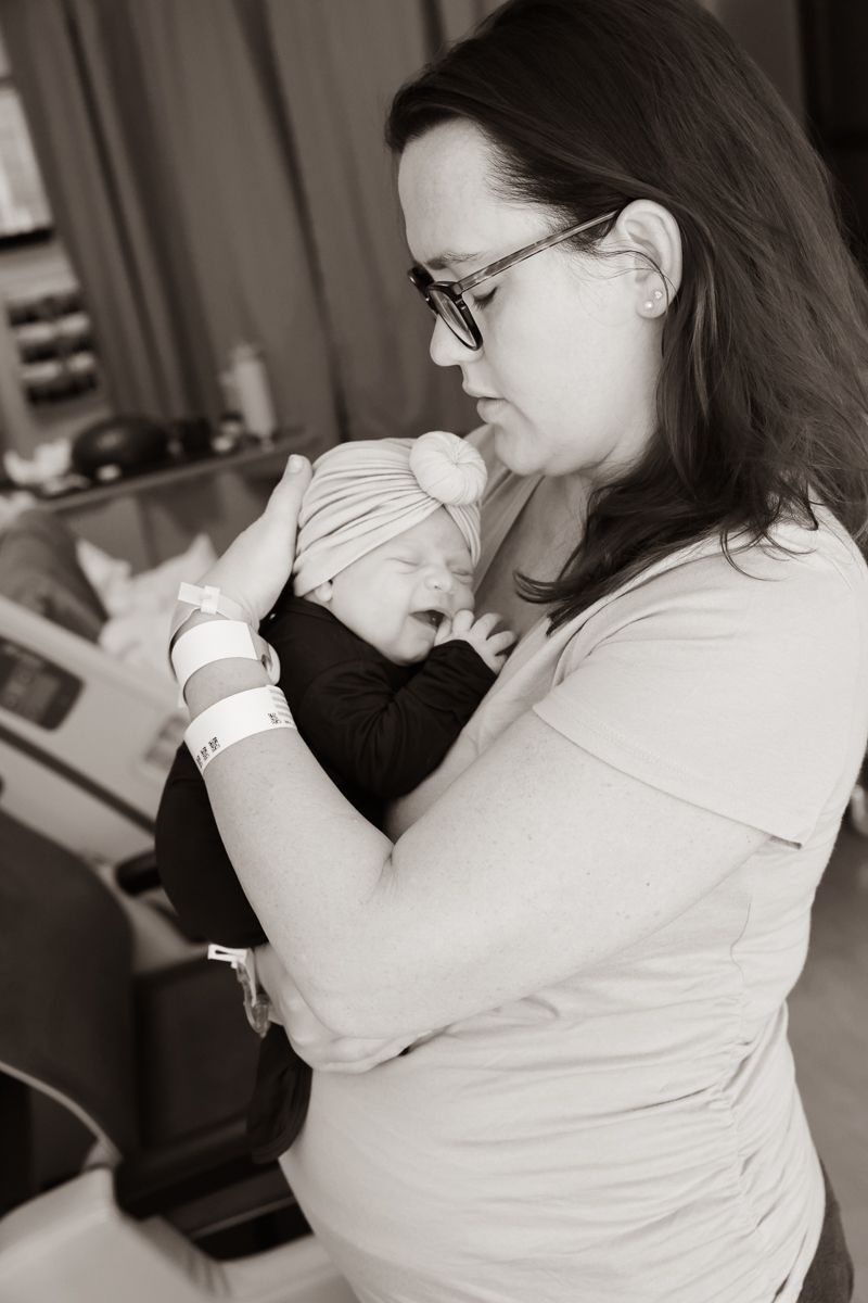 Mother in glasses holds sleeping newborn baby close in monochrome portrait.