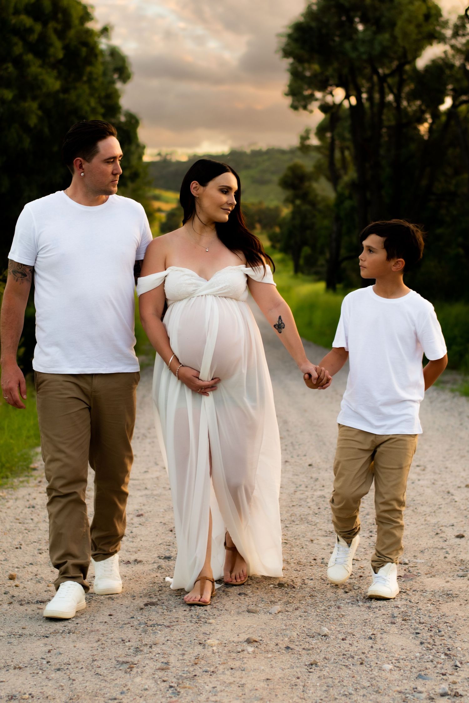 Family of three walking together on dirt path, with expecting mother in white flowing dress.