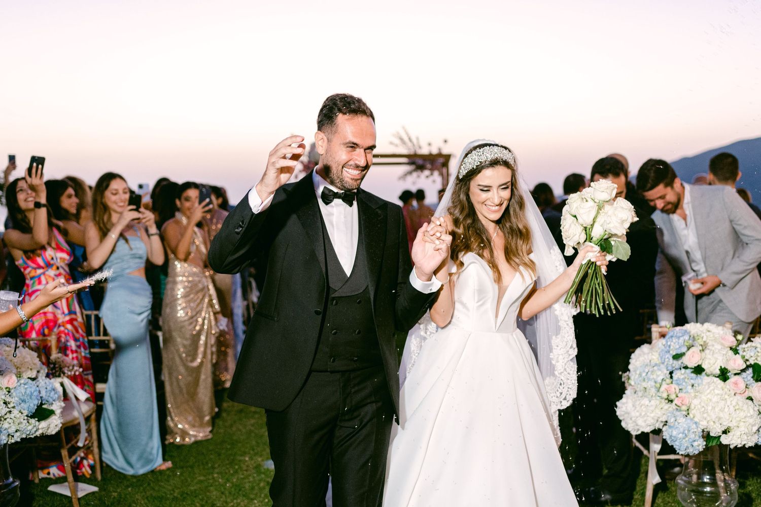 Celebrating couple dancing outdoors at sunset surrounded by guests and floral arrangements.