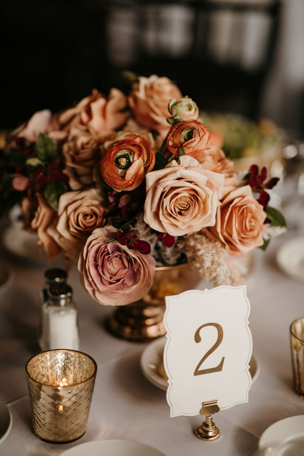 Elegant wedding centerpiece with blush and coral roses in a gold vase beside a table number two card.