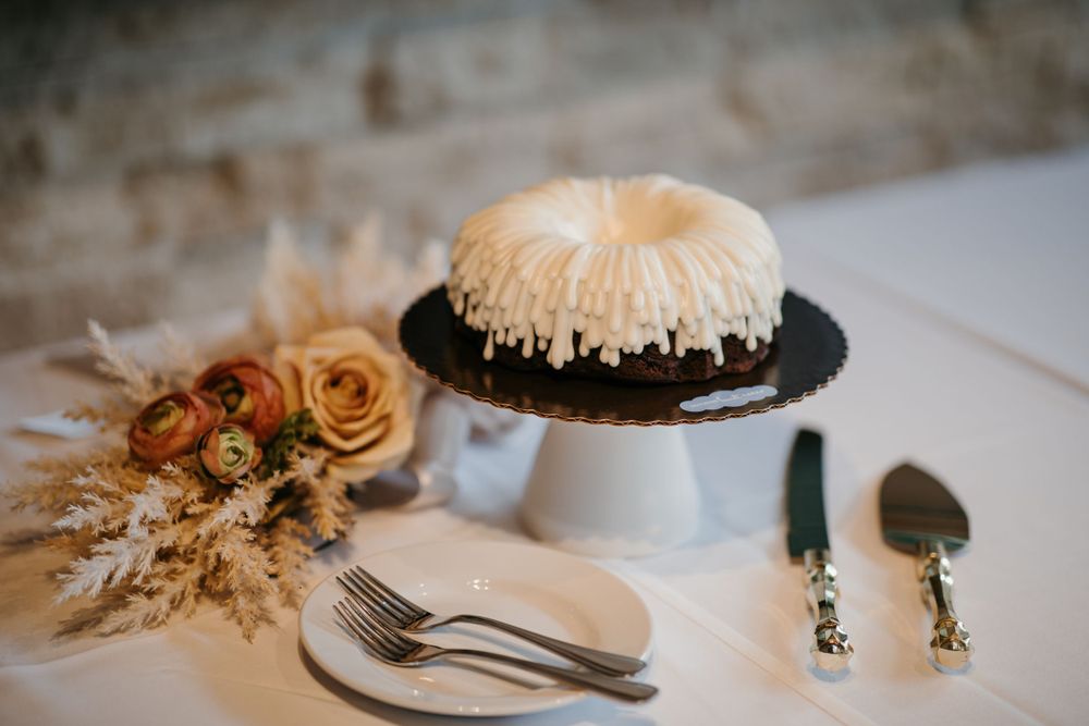 Wedding bundt cake with white frosting on a cake stand beside floral arrangement and serving utensils.