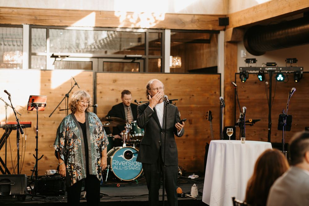 Older couple delivering wedding toast speech with live band and drum kit visible behind them.