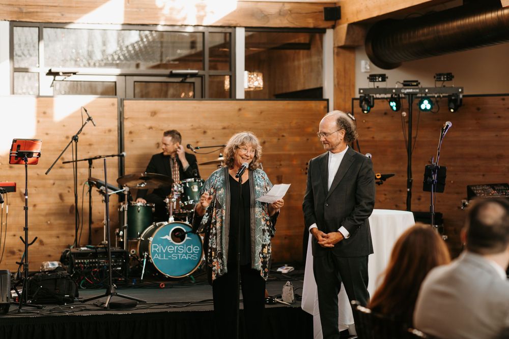 Woman speaking at wedding reception microphone beside man with live band in background.