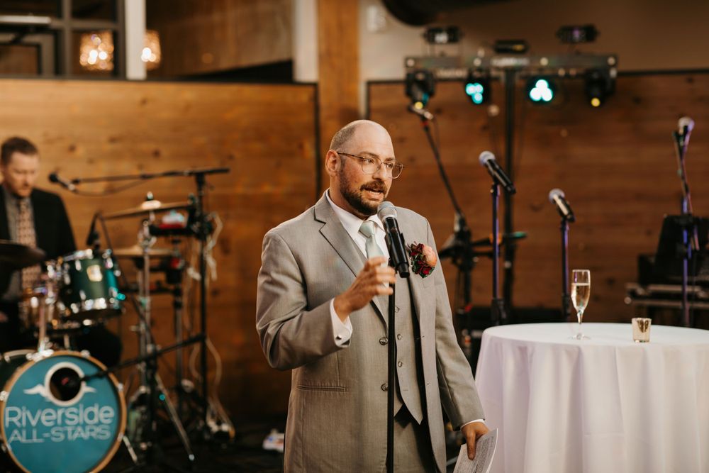 Man in gray suit giving a wedding toast speech with champagne glass on nearby table.