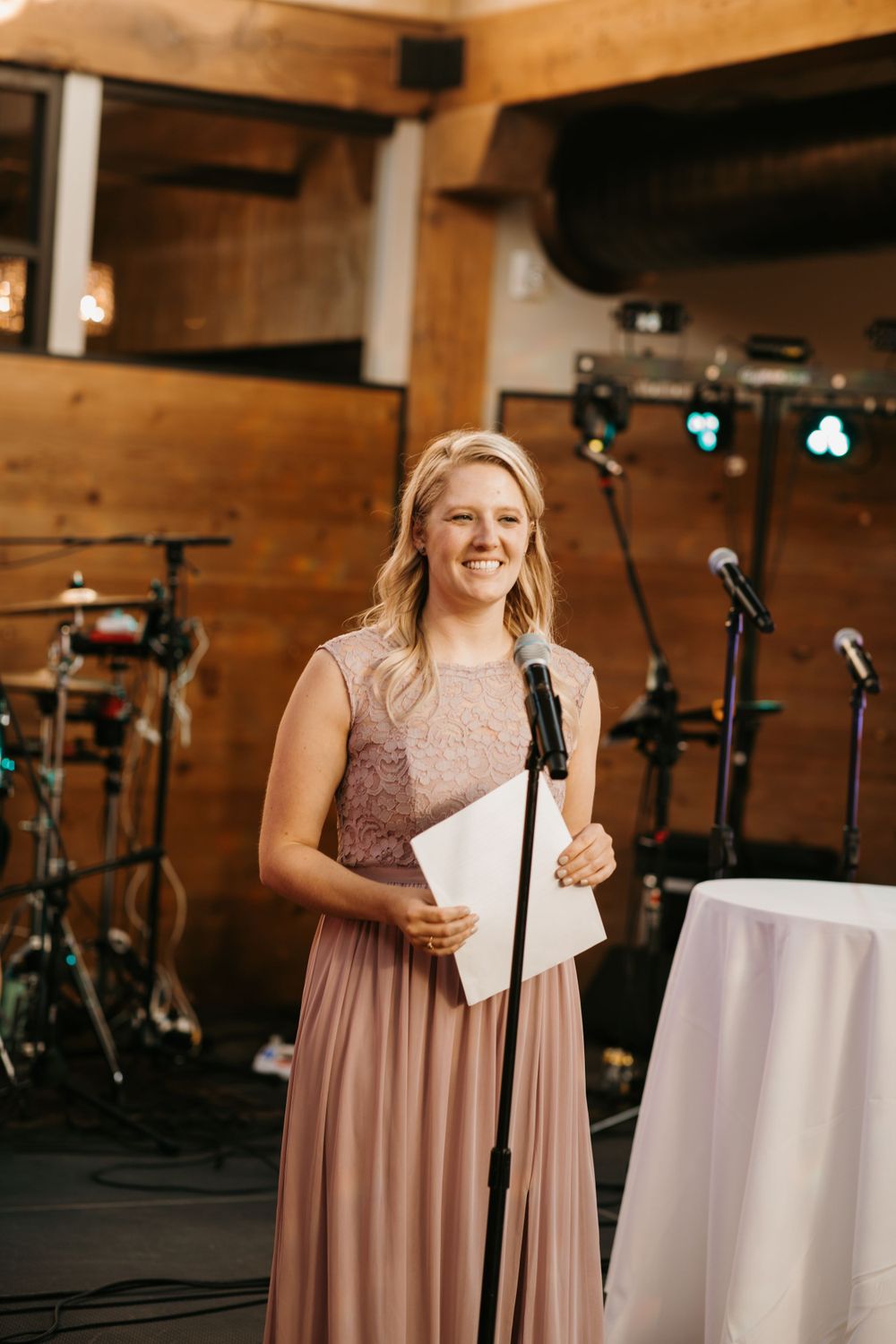 Smiling bridesmaid in dusty rose gown delivers wedding speech at microphone stand in rustic wood venue.