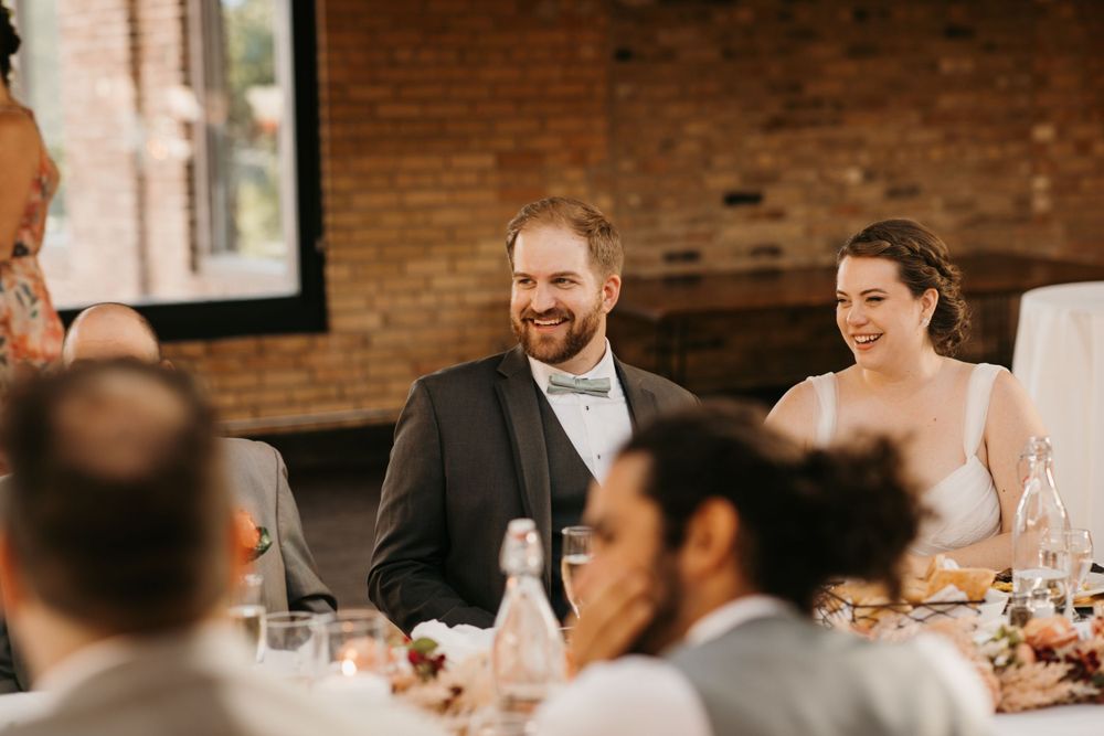 Groom in gray suit and bride laugh joyfully at sweetheart table during wedding reception.