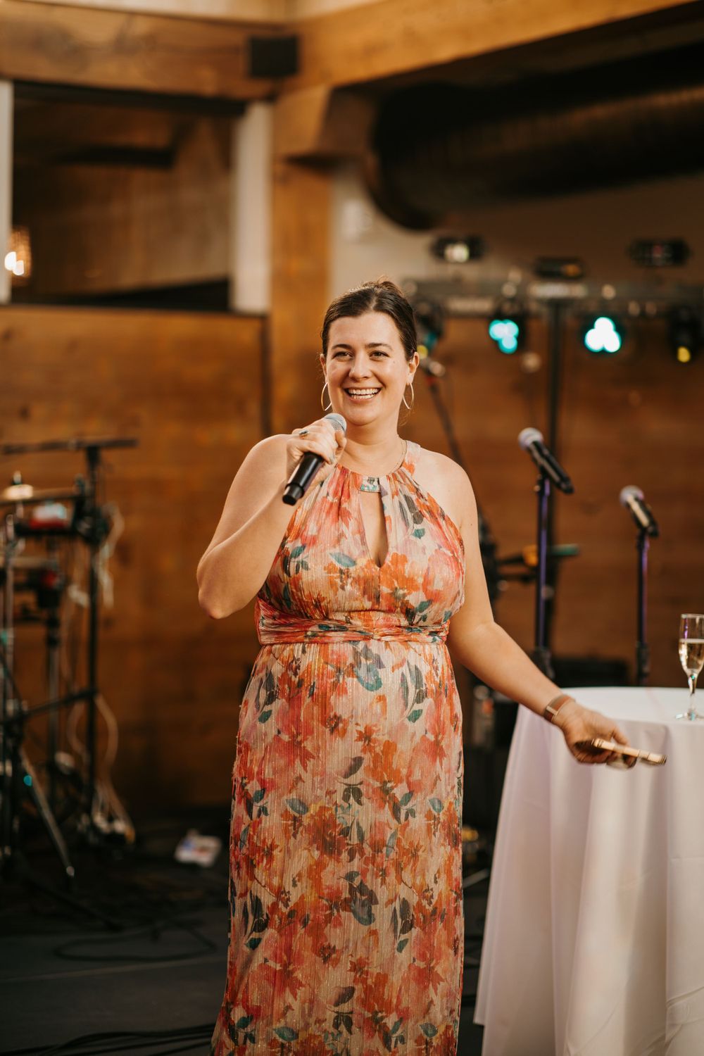 Woman in floral halter maxi dress smiles while giving a toast with microphone at wedding reception.