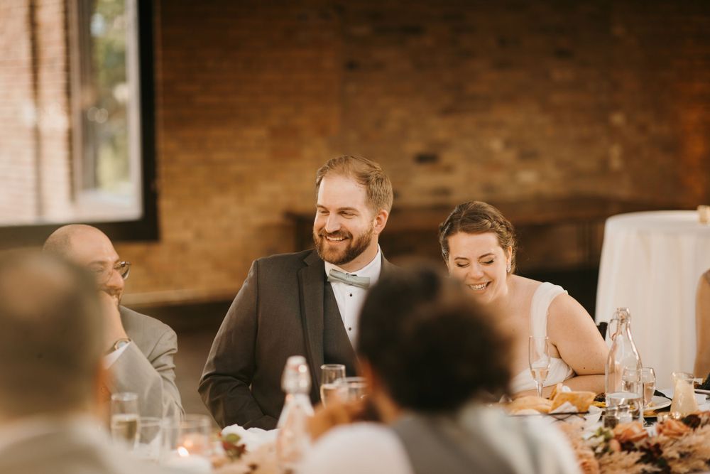 Groom in gray suit and bride share a laugh with guests at elegant wedding reception dinner table.