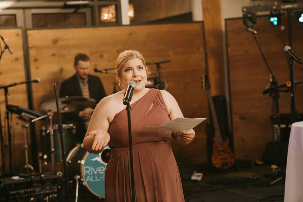 Bridesmaid in mauve halter gown reads heartfelt speech at microphone during rustic barn wedding reception.