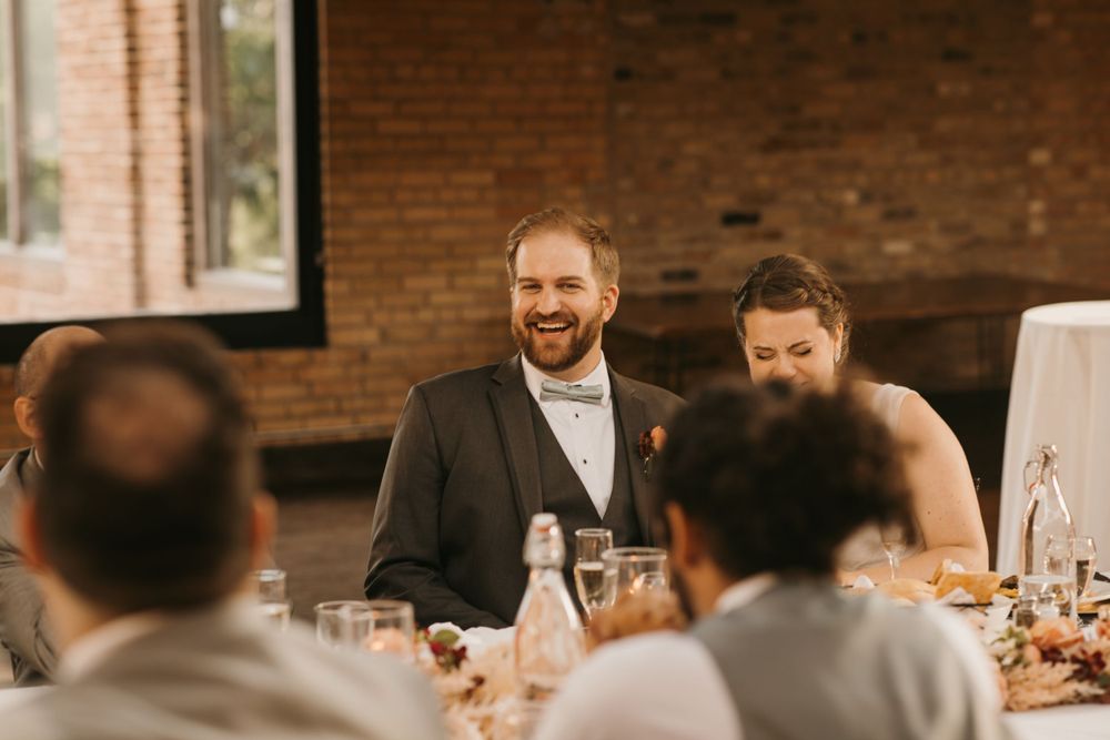 Groom in gray suit with bow tie laughs warmly during toasts at rustic brick wedding reception.