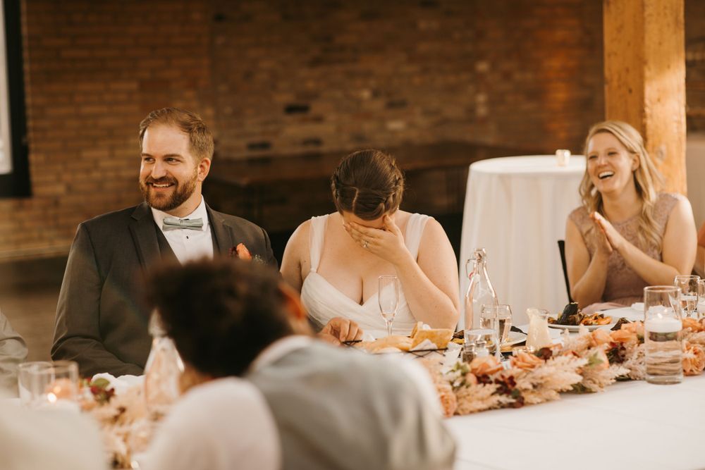 Bride covers her face laughing while groom smiles during funny wedding speech at reception dinner.