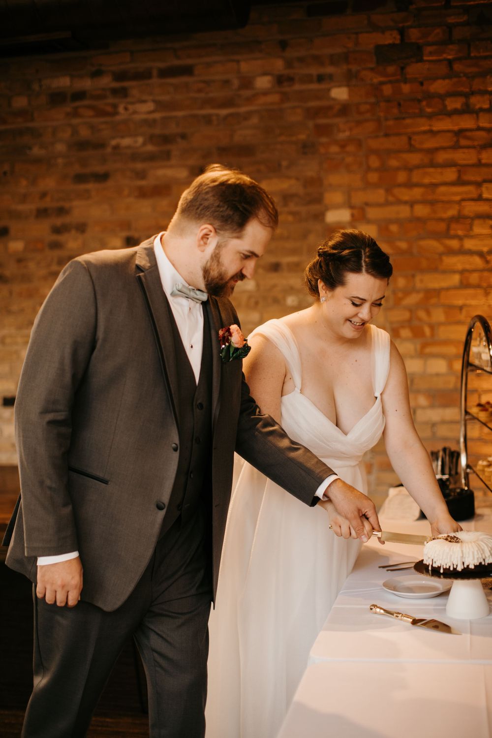Bride and groom smile while cutting their wedding cake together at rustic brick wall venue.