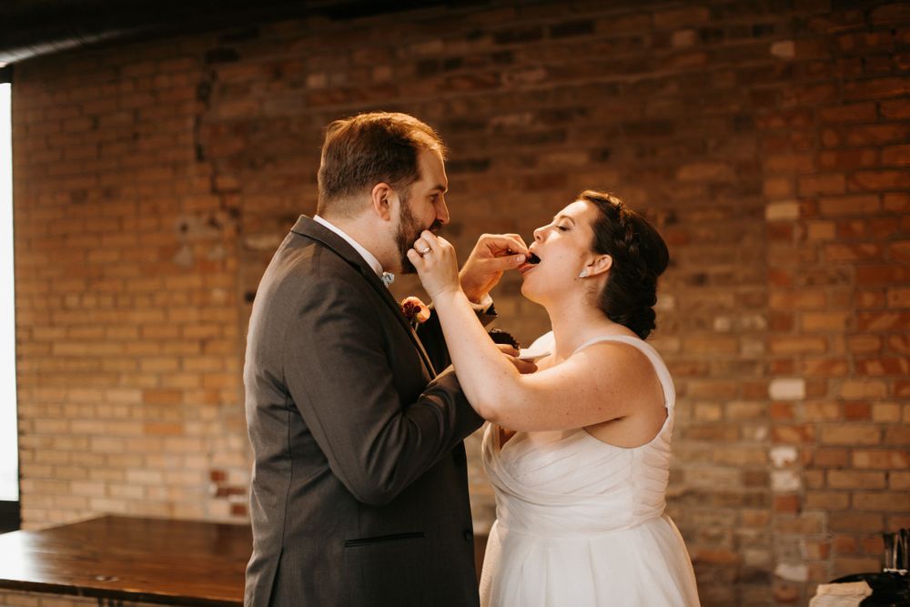 Bride and groom share a joyful cake tasting moment at their wedding reception near a rustic brick wall.