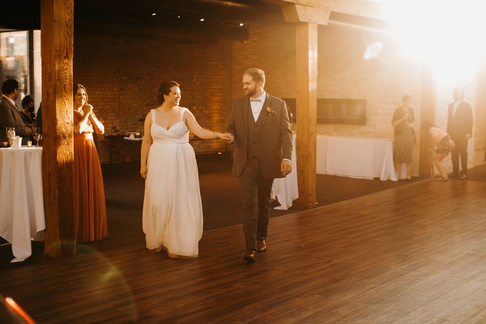 Bride and groom walk hand in hand onto dance floor bathed in golden light at wedding reception.