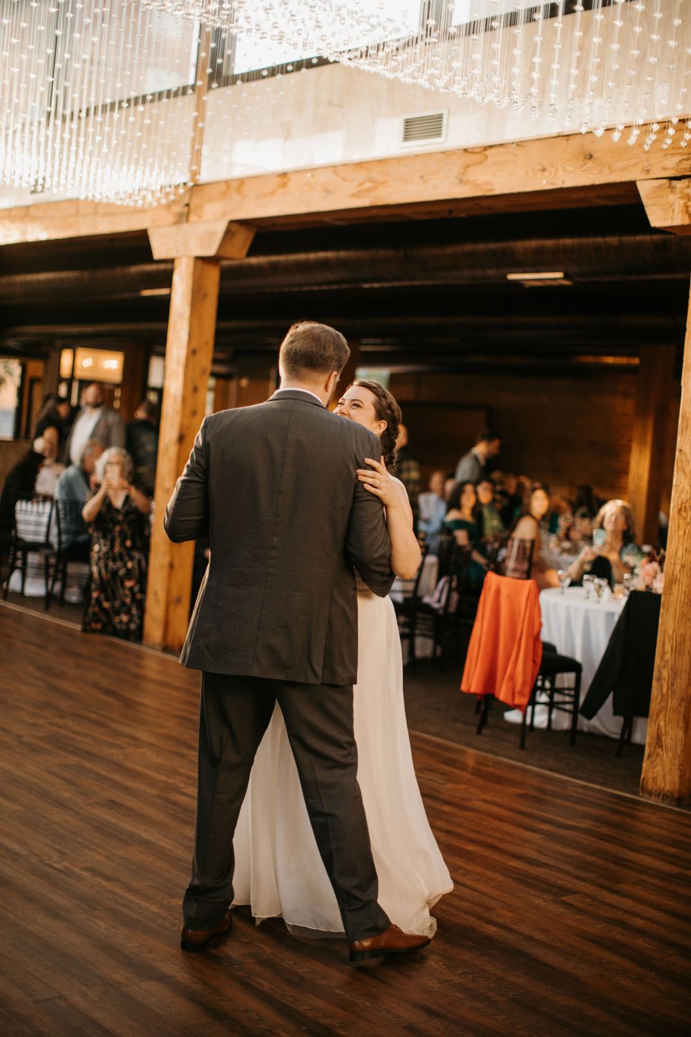 Bride and groom embrace during first dance in elegant rustic venue with crystal curtain backdrop.