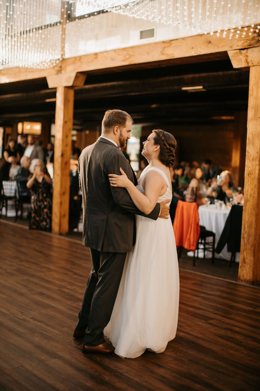 Smiling bride and groom share romantic first dance in warmly lit rustic wedding reception hall.