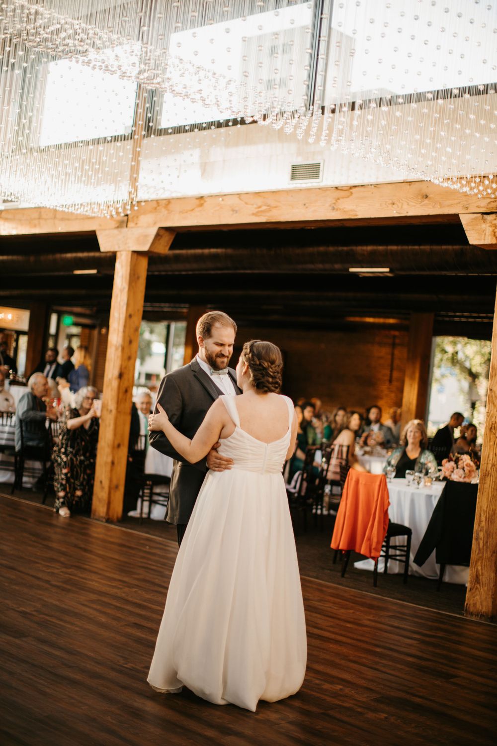 Groom smiles at bride during first dance in rustic venue with sparkling crystal backdrop.