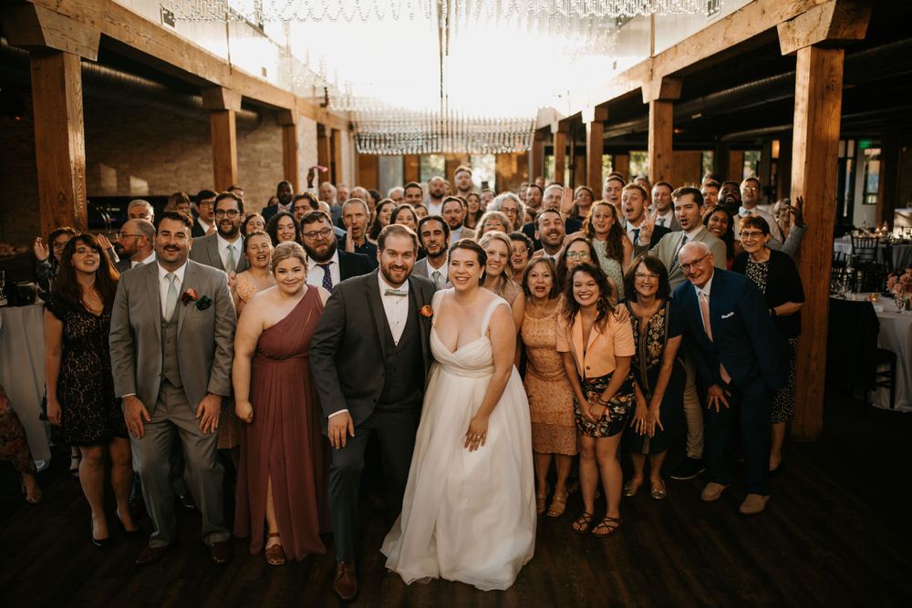 Bride and groom pose with large group of wedding guests in stunning rustic reception venue.