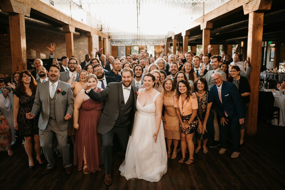 Happy couple celebrates with all wedding guests in large group photo at rustic venue.