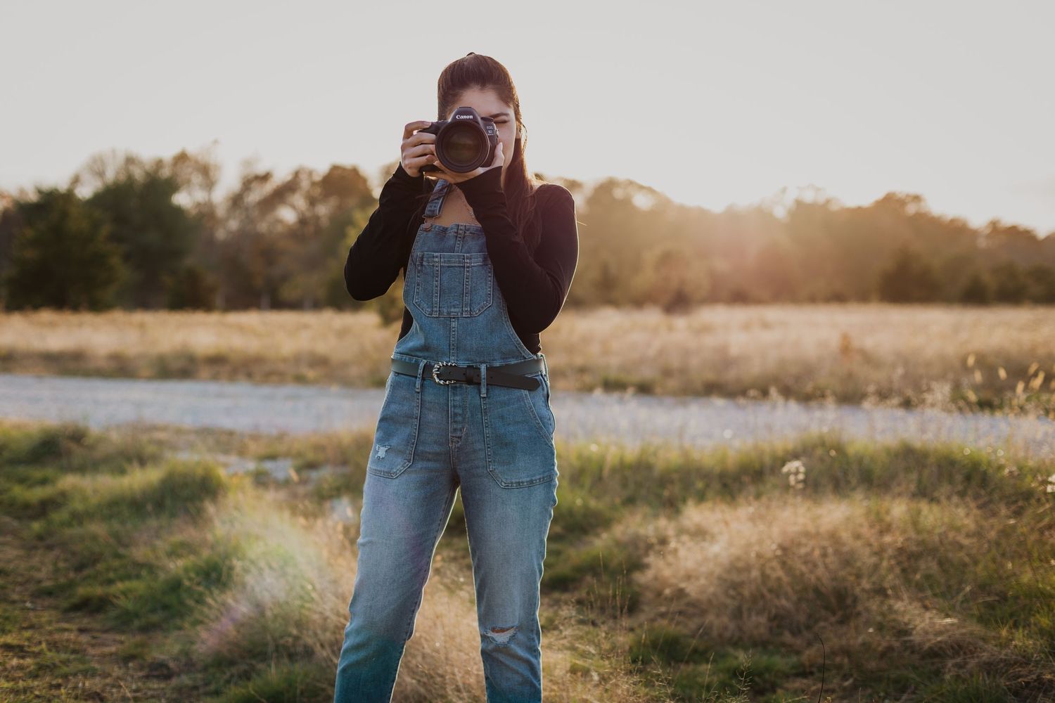 A photographer wearing denim overalls takes pictures in a natural outdoor setting during golden hour.