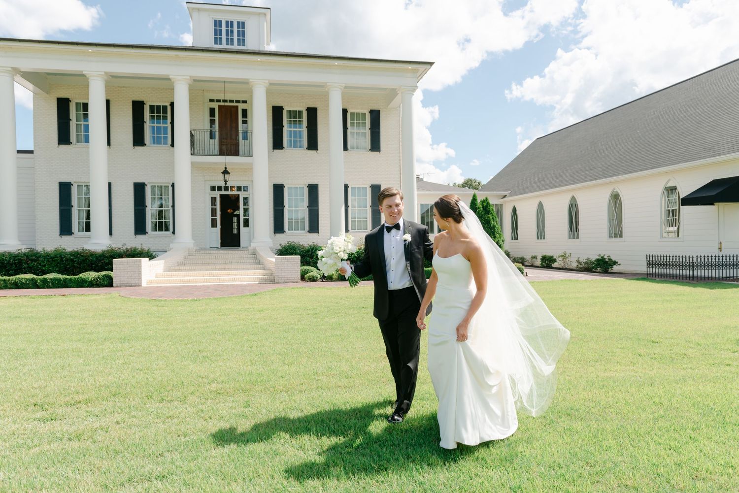 Bride and groom walk hand-in-hand on lush green lawn before elegant white plantation-style wedding venue.