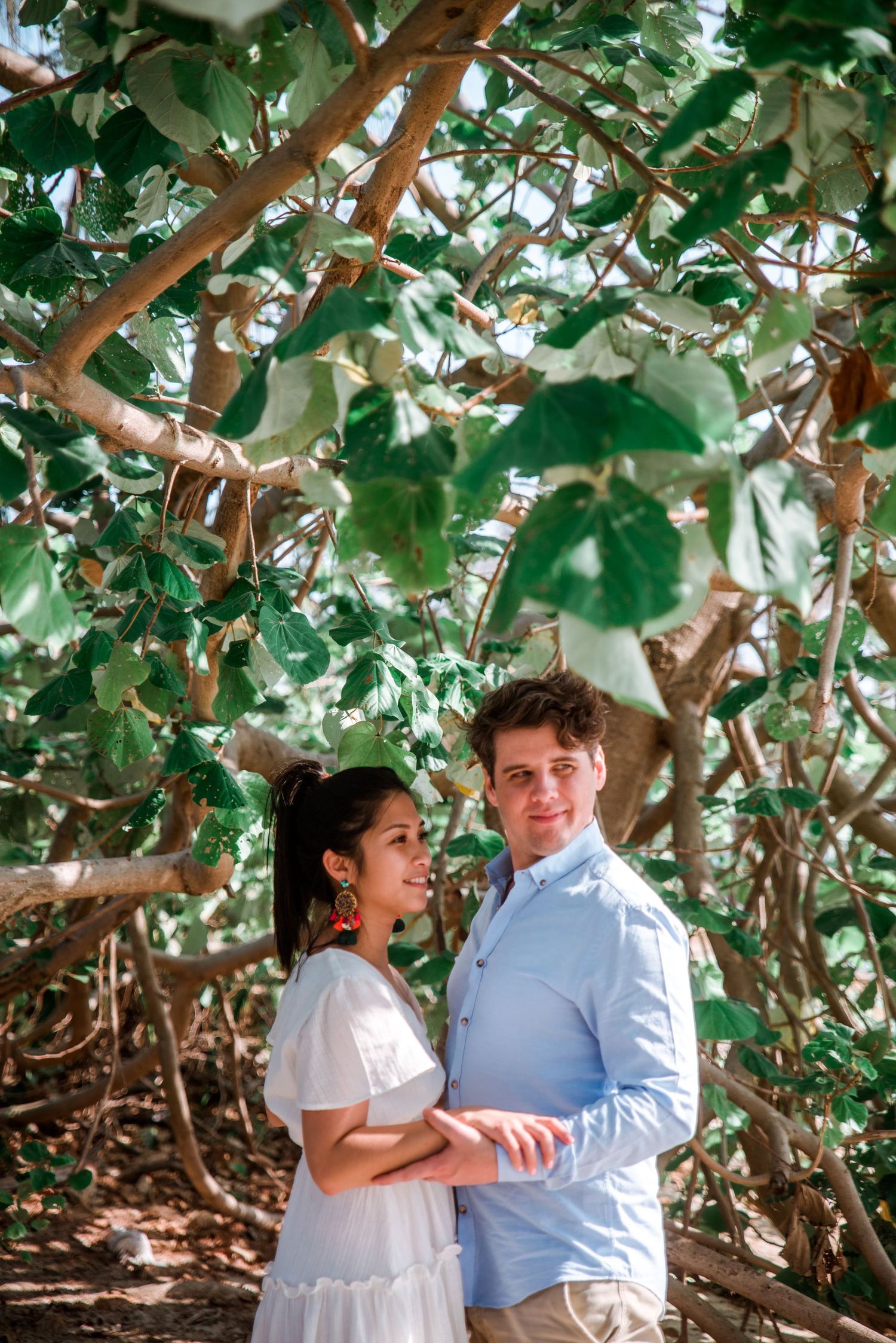 A romantic portrait of a couple standing together surrounded by green leaves and branches.