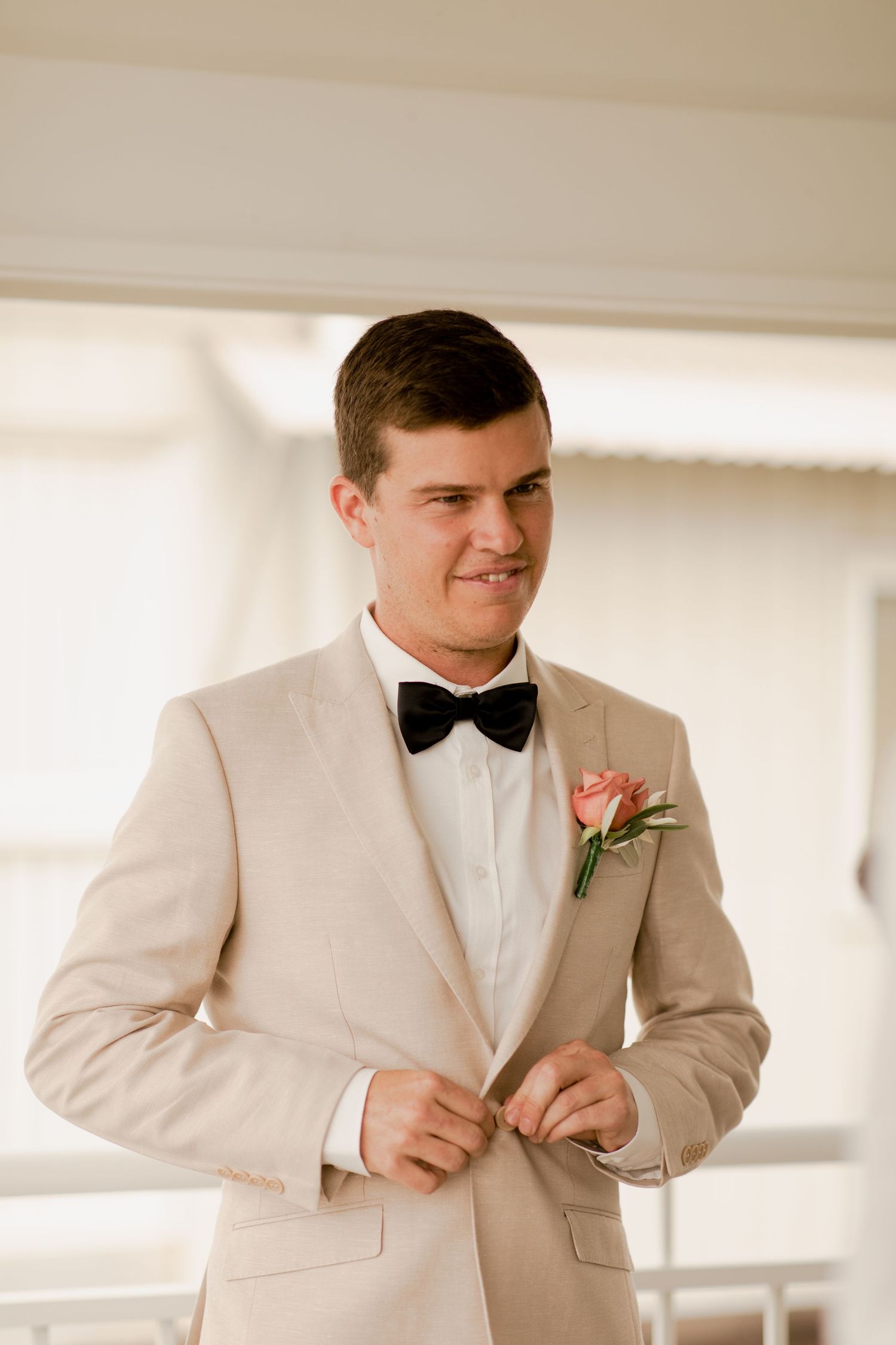 Groom in beige tuxedo with bow tie and coral boutonniere adjusts his cuffs.
