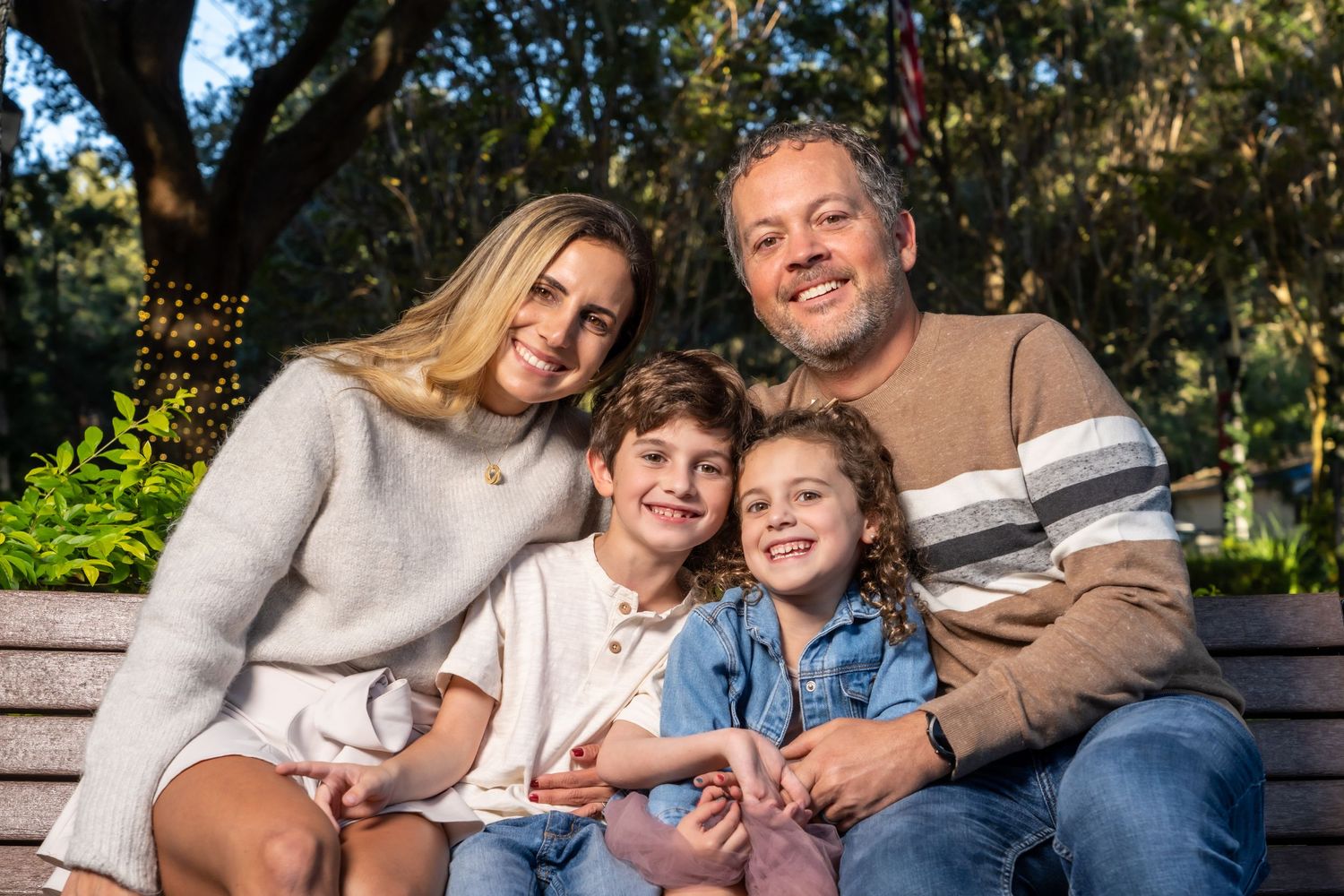 A family shares a close moment while sitting on a wooden bench in a sunlit garden setting.