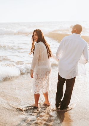 Couple walking along beach shore with waves crashing during golden hour.