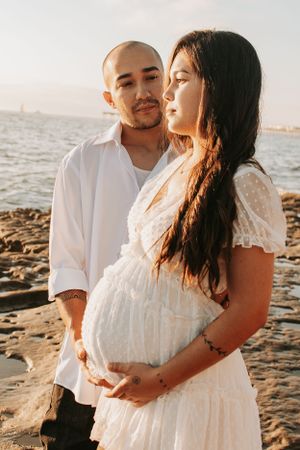 Maternity photo of couple in white clothing on rocky beach at sunset.