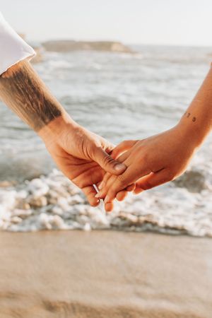 Close-up of two hands clasped together on sandy beach with ocean waves in background.