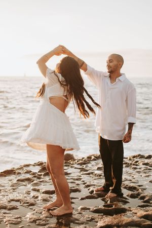 Playful beach photoshoot with couple in white outfits on rocky shore.
