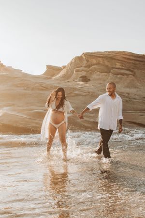 A couple splashes through shallow water at sunset near rocky cliffs.