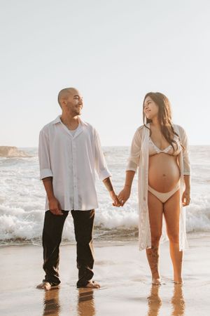 A couple holds hands while standing at the beach edge during sunset.