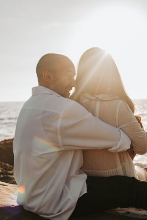 A couple embraces while sitting on coastal rocks during golden hour sunset.