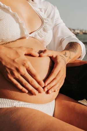 Close-up of hands tenderly cradling pregnant belly in white lace dress.