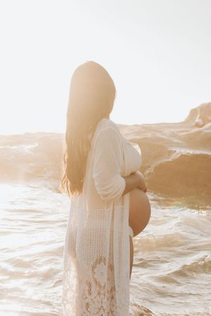 Silhouette of expecting mother in white lace dress standing by ocean at sunset.