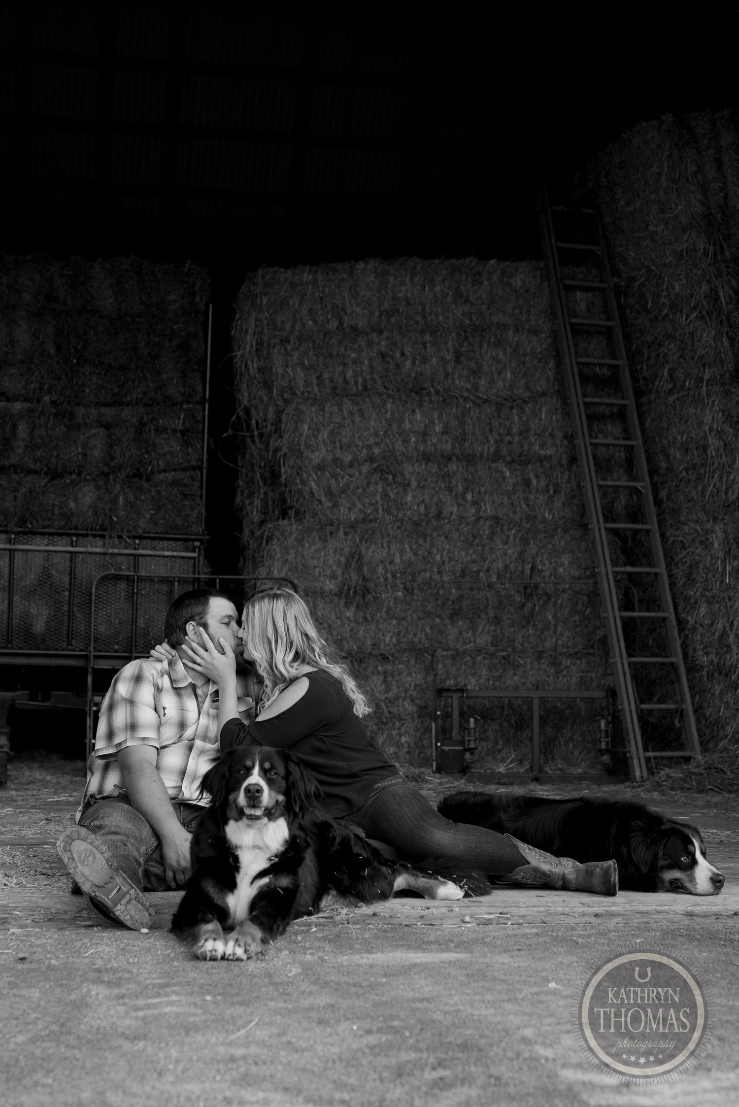 Romantic black and white photo of a couple with their dog in a rustic barn setting.