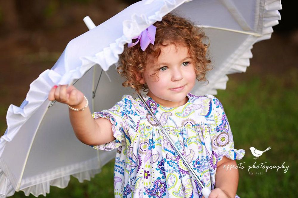 Child holding white lace parasol wearing floral dress in outdoor portrait.