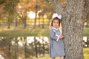 A child in a gray dress standing by a large tree trunk near a pond at sunset.