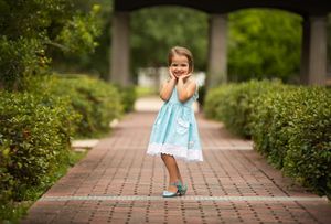 A child in a light blue dress standing on a brick pathway surrounded by hedges.