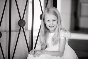 A black and white portrait of a child in white lace sitting by a decorative iron railing.