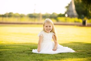 Child in white dress sitting on sunlit grassy field with church steeple in background.