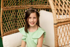 Portrait in mint green flutter sleeve dress seated in wicker peacock chair.