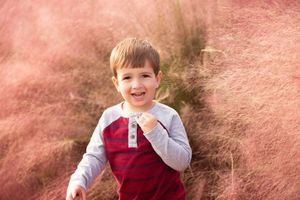 Child wearing red vest and gray shirt poses against pink background with ornamental grass.
