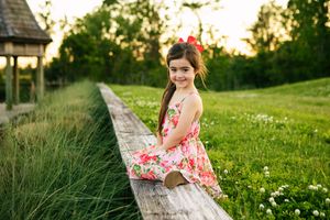 Person in floral dress poses on wooden fence during sunset in garden setting.